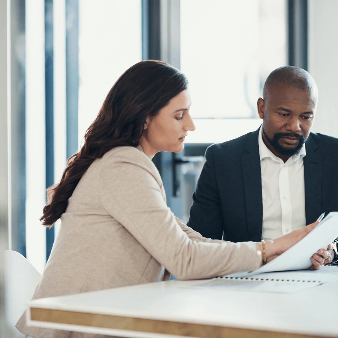 Two people in business attire sit at a table, reviewing documents together in an office setting