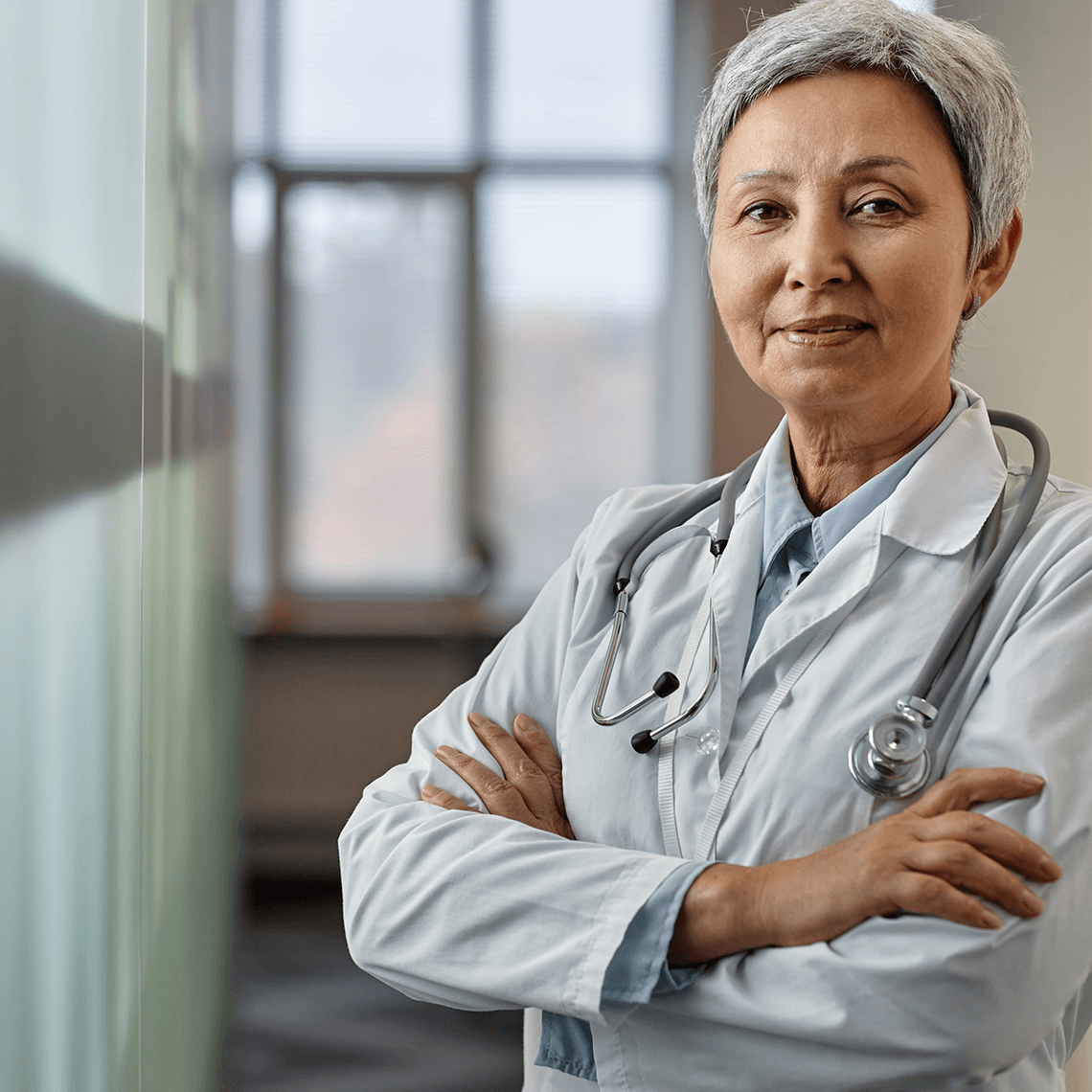 A woman in a doctor's coat stands by a window, looking confidently at the camera