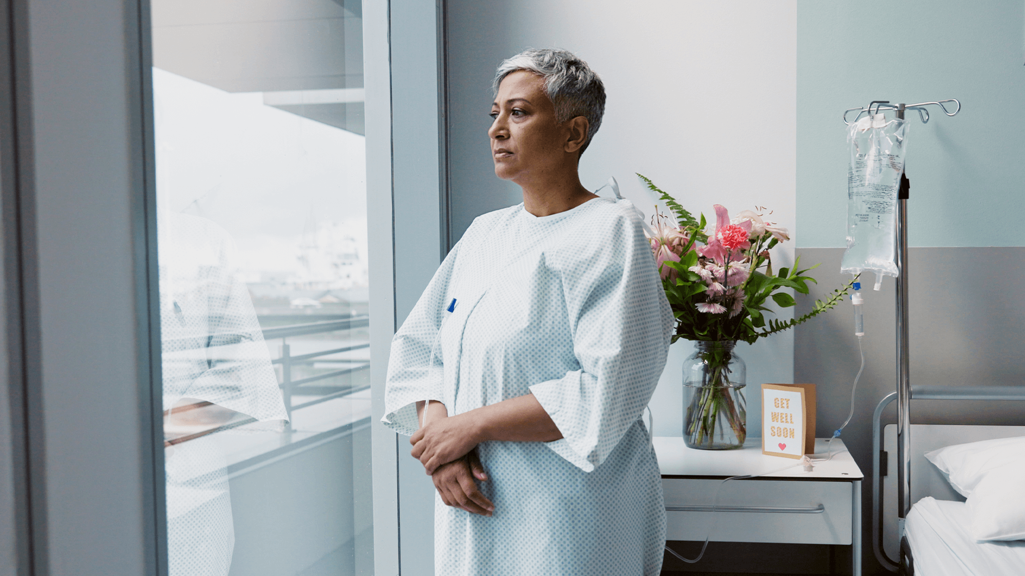 A patient in a hospital gown stands by a window, gazing outside. A colorful bouquet and a "Get well soon" sign adorn the bedside table.