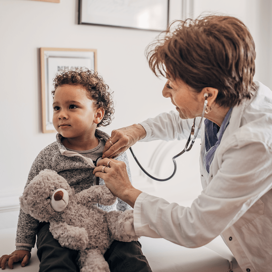 Little curly-haired boy with teddy bear gets heartbeat measured by female doctor