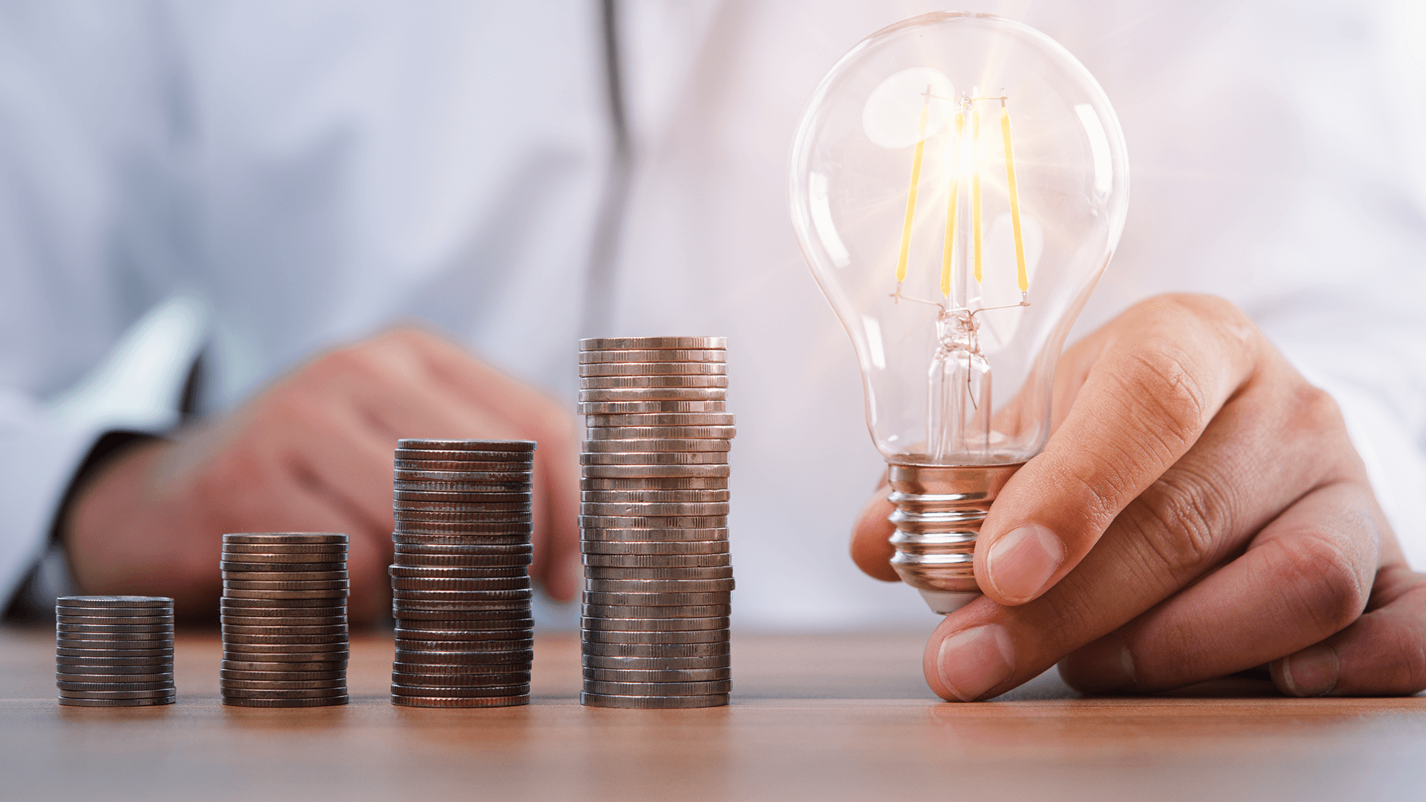 A person holding a glowing light bulb in one hand, while the other hand points at stacks of coins, symbolizing financial ideas or investments.