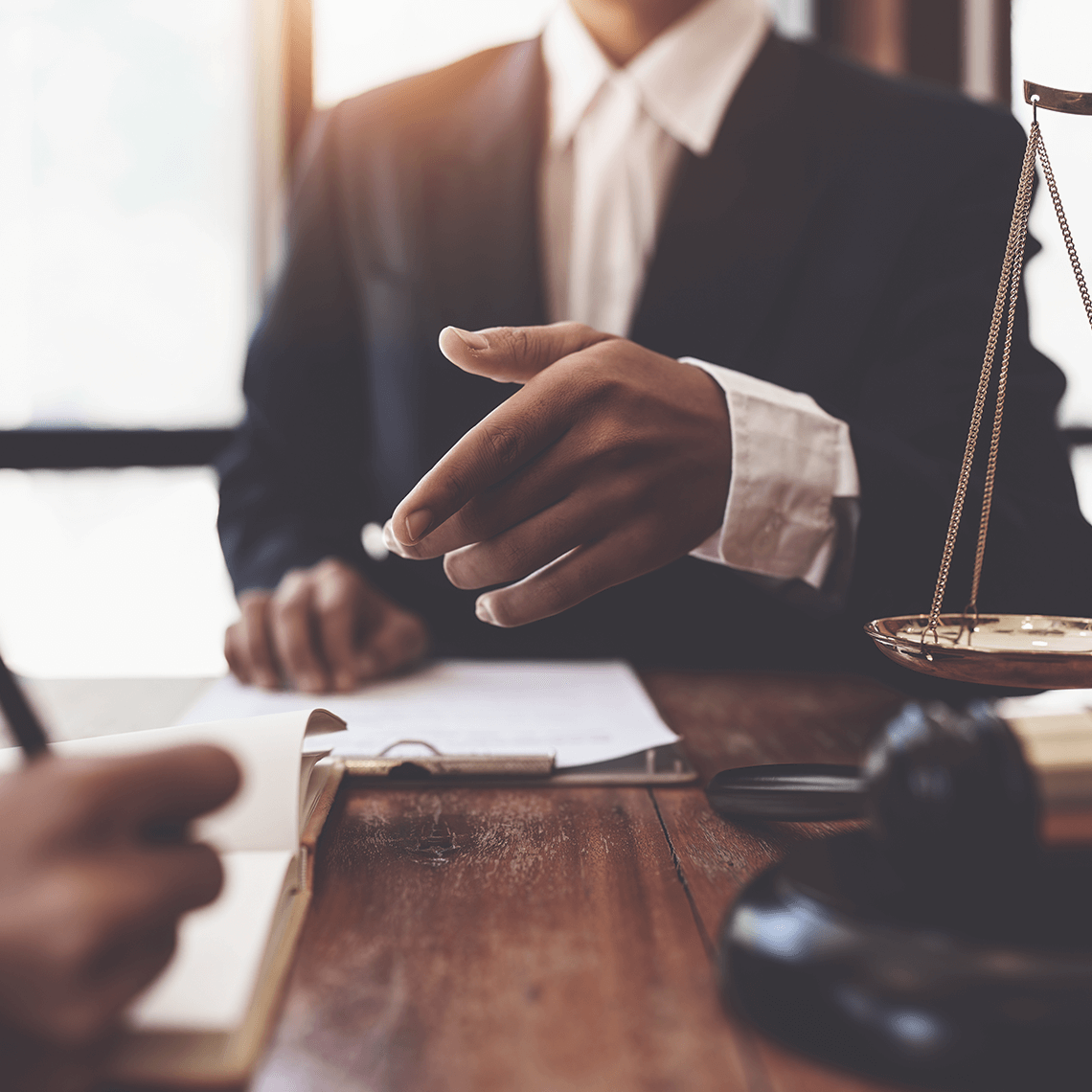 Two people meet at a desk with legal documents, a gavel, and scales of justice, suggesting a legal or judicial discussion.