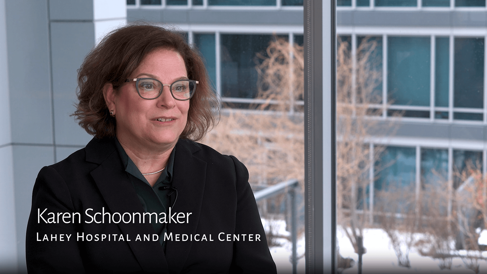 A woman with glasses and curly hair smiles while speaking, seated indoors with a snowy window view behind her. She wears a black blazer, creating a professional tone
